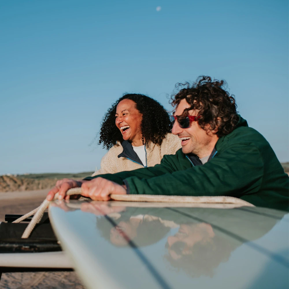 Couple on beach laughing leaning on a surf board